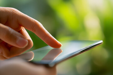 Person using a smartphone with a finger tapping on the screen, reflecting sunlight with a blurred natural green background in soft focus