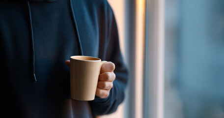 Person wearing dark hoodie holding a beige coffee mug near a window with soft natural light and blurred background