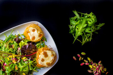 Gourmet Salad with Baked Cheese Wrapped in Kataifi Pastry, Mixed Greens, and Scattered Pistachios on a Black Background
