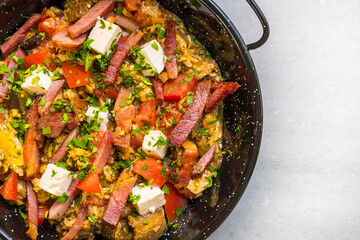 Hearty Traditional Greek Omelet or Fry-Up with Scrambled Eggs, Feta Cheese Cubes, Sliced Cured Meat, and Tomatoes in a Black Skillet