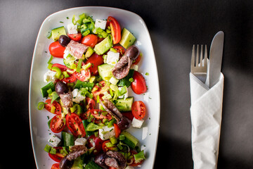 Overhead Tabletop View of Traditional Greek Salad with Feta Cheese, Olives, Cured Sausage, and Wrapped Cutlery