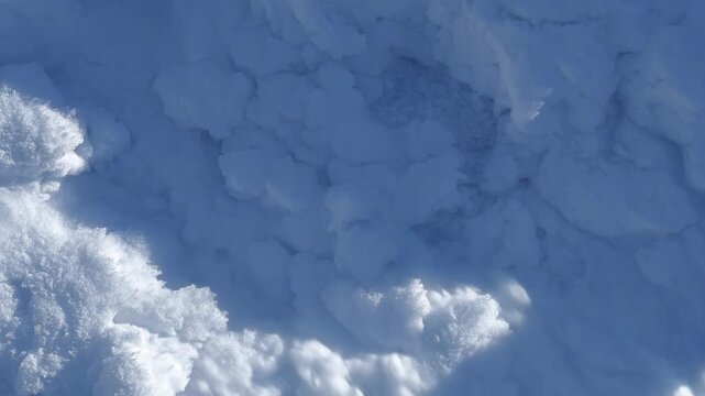 Bright sunlight casting a moving shadow over a pristine, bumpy texture of fresh, white snow. Close-up detail of winter ground cover