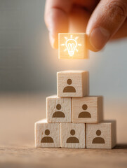 Wooden blocks arranged in a pyramid with human icons and a glowing light bulb symbolizing leadership and innovative ideas being placed on top by a hand