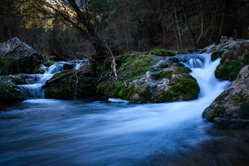 R&iacute;o de monta&ntilde;a con efecto seda en el agua fluyendo sobre rocas cubiertas de musgo en un bosque oscuro.