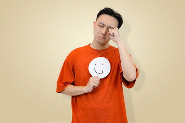 Distressed young Asian man in an orange t-shirt rubbing his eye as if crying or in pain, while holding a happy face emoji sign, symbolizing the effort to mask sadness or exhaustion 