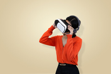 Amazed young Asian woman in orange clothing wearing a virtual reality headset and headphones, looking and holding the device, symbolizing the thrilling discovery in studio background