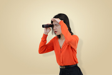 Fototapeta premium Intense young Asian woman in orange clothing looking through a monocular symbolizing focused observation, careful search in studio background