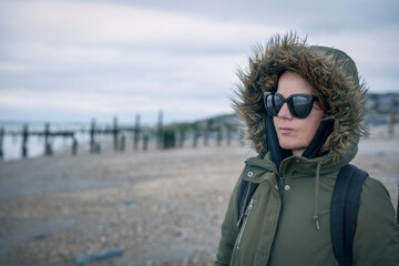 Woman in fur hooded winter coat with sunglasses and backpack on windy beach