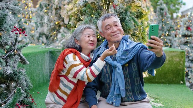 In a cozy outdoor setting, an elderly asian couple shares joyful moments beneath a beautifully decorated Christmas tree, snapping selfies and admiring their cheerful photos together in the winter