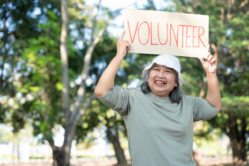 A senior woman smiles while holding a VOLUNTEER sign in a sunny park. The image highlights active aging, community service, and the spirit of giving back through outdoor social engagement