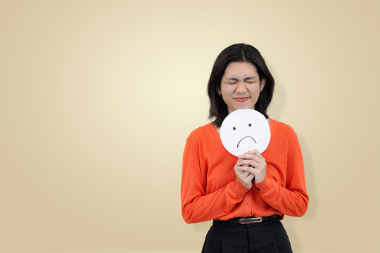 young Asian woman in an orange sweater holding a sad face emoji sign to her chest with eyes closed, clearly expressing sadness, distress, or emotional suffering without masking her true feeling - Powered by Adobe