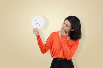 Smiling young Asian woman in an orange sweater, holding a sad face emoji sign to partially cover...