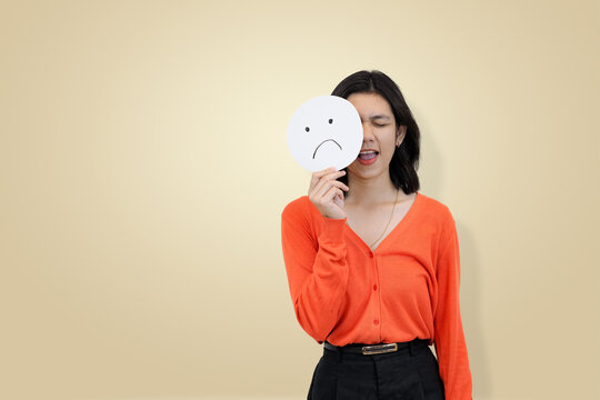 Smiling young Asian woman in an orange sweater, holding a sad face emoji sign to partially cover her face, symbolizing the emotional contradiction of masking inner sadness - Powered by Adobe