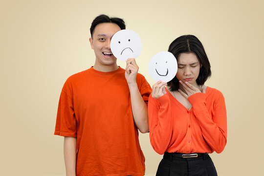 Young Asian man and woman in orange tops standing together, holding paper circles with contrasting sad and happy face emojis over their own faces, symbolizing emotions, hidden feelings