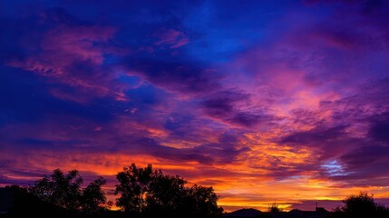 Obraz premium The sky transforms into a canvas of brilliant hues as the sun sets behind silhouettes of trees.