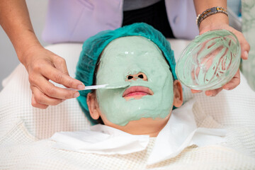 Asian woman receiving professional facial treatment at beauty spa salon. Beautician applying green...