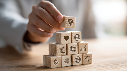 a person stacking wooden cubes into a pyramid shape, each cube has an icon: heart, gear, graph, globe, on a light wood table, natural daylight