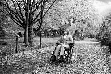 Elderly woman with grey hair in wheelchair on a walk in the park in fall weather with nurse or health care worker in blue uniform, both smiling happy