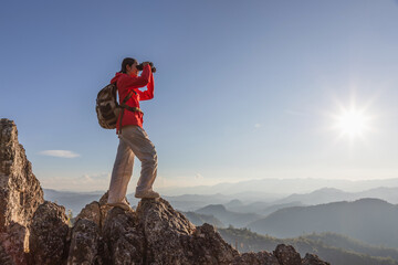 Naklejka premium Cute girl hiker with backpack holding binoculars standing on top of the rock mountain sunset sky background