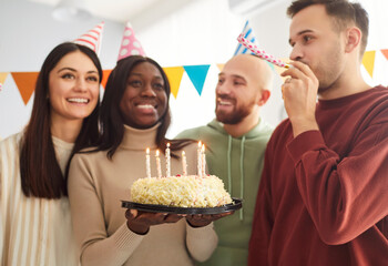Group of cheerful optimistic friends having fun on birthday party, congratulate their best friend with anniversary, blowing in party horns and going to blow candles on cake. Celebration concept