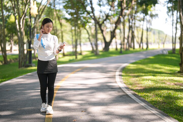 Young Asian woman prepares for outdoor workout in a green park adjusting wireless earbuds with her smartphone promoting health and wellness