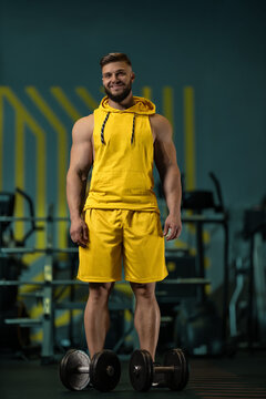 man stands in a gym wearing yellow workout clothes with a smile. He is positioned next to a set of dumbbells and looks ready for exercise.
