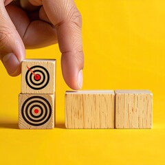 Hand placing a wooden block with target image on top of stacked blocks on yellow background