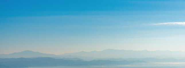 Fototapeta premium Minimalistic landscape of blue mountain layers in soft morning mist and haze. Tranquil panoramic natural background with a gentle color gradient and vast copy space for design.