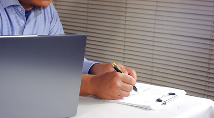 Closeup of a hand holding a pen writing, signing a document with a blurred background, a laptop...