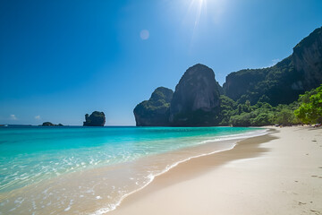 A stunning tropical beach scene featuring pristine white sand, crystal-clear turquoise ocean water, and gentle waves. The background is dominated by dramatic, steep limestone (karst) cliffs under a br