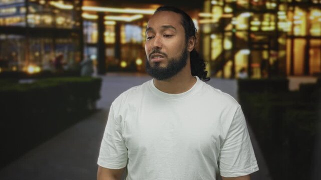 Man touching forehead with hand outside a lit building on a city street at night, wearing a white tshirt; frustration.