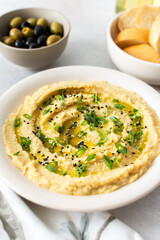 A bowl of creamy hummus topped with olive oil and herbs sits beside fresh bread and a bowl of olives. The light setting showcases a simple, inviting meal