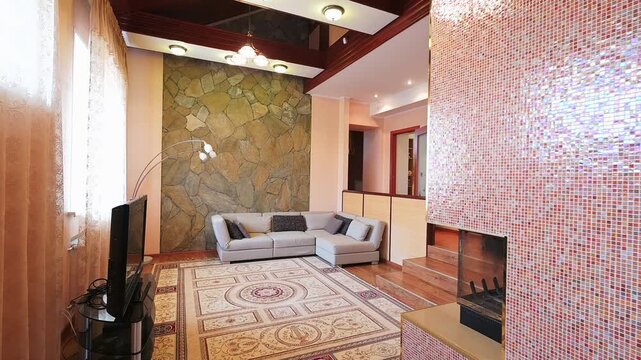 A cozy living room with a stone accent wall, L-shaped sofa, patterned rug, wooden ceiling beams, chandelier, TV stand, and open shelving. Warm wood tones and soft lighting create a welcoming atmospher