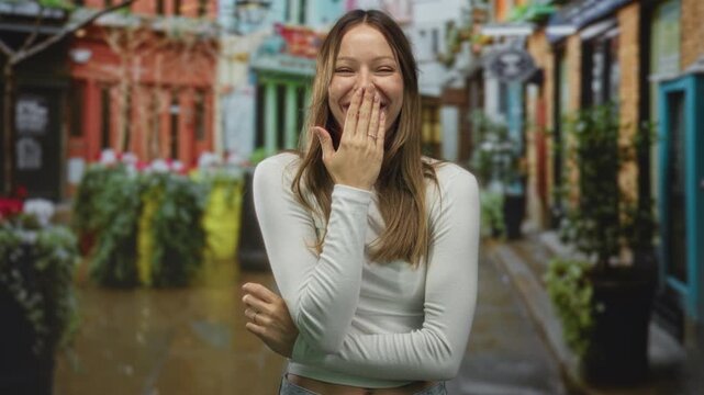 Woman covering mouth with hand on a city street while smiling and crossing her arms, casual urban scene; surprise candid moment.