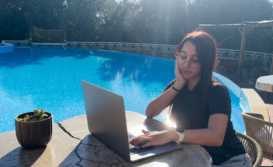 Freelancer using laptop working remotely near swimming pool. Young traveler girl working on computer during her summer holiday. Work from anywhere, Technology and lifestyle concept, copy space