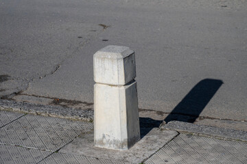 Naklejka premium A concrete bollard casts a long shadow on a patterned sidewalk beside a cracked urban road.
