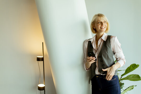 Female professional with short hair stands by wall while using smartphone in modern office
