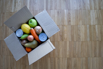 Box with assorted groceries on a wooden floor. Food donation package with fresh fruits, vegetables, and canned goods. Top view of an open cardboard box filled with mixed produce.