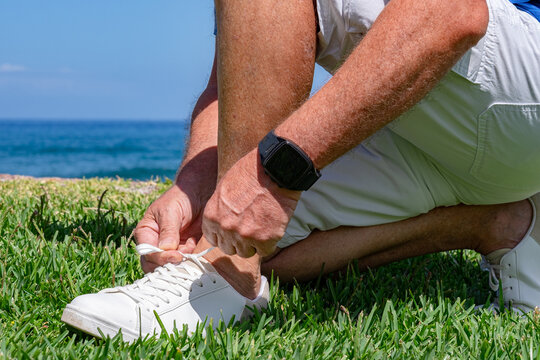 Close-up of a senior man kneeling on the grass and tying the laces of his white sneakers near the ocean on a sunny day. Active lifestyle, outdoor leisure, and positive atmosphere - Powered by Adobe
