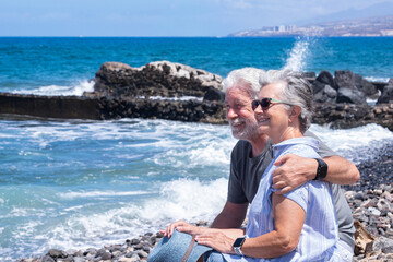 Cheerful relaxed senior couple hugging and smiling enjoying a peaceful moment by the seaside sitting near the beach with waves and clear blue sky in the background