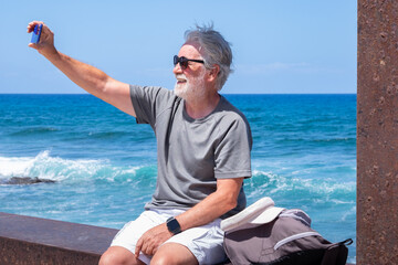 Senior man smiling taking a selfie by smartphone enjoying a peaceful moment by the seaside sitting near the beach with waves and clear blue sky in the background. Relaxation and retirement