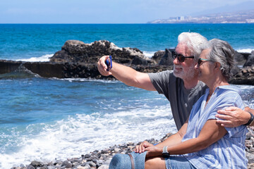 Cheerful relaxed senior couple hugging and smiling taking a selfie by smartphone enjoying a peaceful moment by the seaside sitting near the beach with waves and clear blue sky in the background