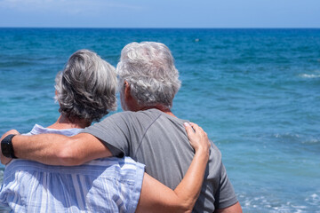 Relaxed senior couple hugging  looking at the horizon enjoying a peaceful moment by the seaside sitting near the beach with waves and clear blue sky in the background. Retirement concept