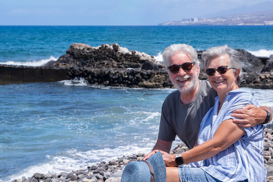Cheerful relaxed senior couple hugging and smiling looking at camera enjoying a peaceful moment by the seaside sitting near the beach with waves and clear blue sky in the background - Powered by Adobe