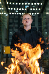 A man in a black hoodie stands centered, illuminated by the intense flames of a bonfire in the foreground