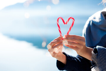 Two candy canes in shape of heart in woman hands. Christmas mood, happy Valentine's day. Concept of love, winter holidays, sweetness, warmth, festive moments. Close up. Outdoor nature background
