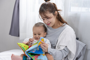 Asian Young mother reading colorful fabric book to baby on bed, tender moment of bonding and early learning through play