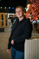 Smiling man in a black hoodie and jeans leans casually against a light wooden planter