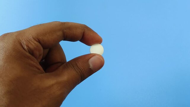 Closeup of asian or indian person hand holding round white medicine pill with finger isolated on blue background and copy space. Painkiller, antibiotic and antidepressant drug bad side effect concept.