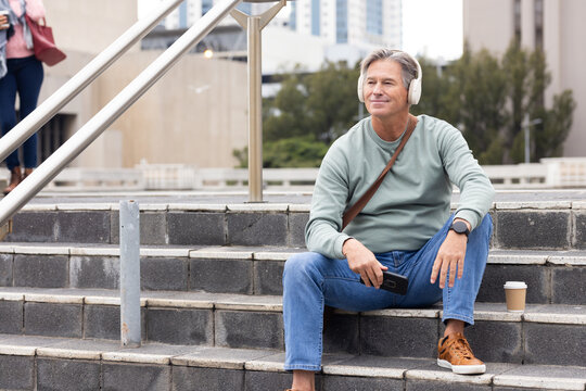 Senior man sitting on plaza steps wearing headphones using smartphone beside coffee cup, copy space - Powered by Adobe